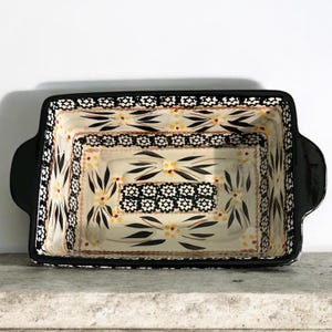 May include: Rectangular ceramic baking dish with black handles and rim. The interior features a cream-colored background with a floral pattern in black, yellow, and brown. The dish is designed for baking and serving.