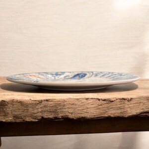 May include: A ceramic plate with a blue and white decorative pattern. The plate is round and has a slightly raised edge. The design includes swirls and floral elements. The plate is resting on a wooden surface.