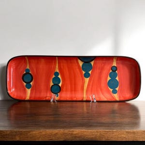 May include: Rectangular ceramic serving platter with a vibrant red background and black trim. The platter features a unique design of teal and yellow abstract shapes. The serving dish is displayed on a wooden surface.