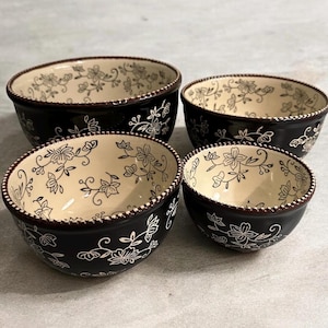 May include: Set of four ceramic bowls in varying sizes. The bowls have a black exterior with a cream-colored interior. Each bowl features a white floral pattern and a decorative rim.
