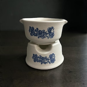 May include: A cream-colored ceramic bowl with a blue floral pattern sits atop a matching cream-colored ceramic base with a corresponding blue floral design. The bowl features a wide, open top with small pouring spouts on opposite sides.