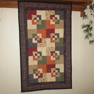 May include: A rectangular quilt with a patchwork design, featuring squares in various colors like red, orange, green, and cream. The quilt has a dark border with a swirling pattern and is hanging on a wall.