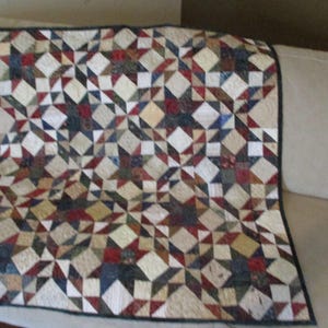 May include: A rectangular quilt featuring a repeating star pattern composed of small fabric squares and triangles. The design incorporates a palette of cream, deep red, navy blue, and muted green tones, finished with a dark solid border.