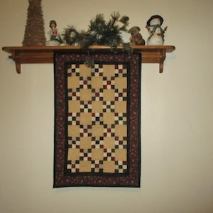 May include: A quilt with a black, white, and red checkered pattern hangs on a wall above a wooden shelf. The shelf is decorated with a small artificial Christmas tree, a snowman figurine, and a small dog figurine.