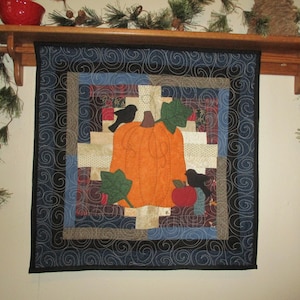 May include: A square fabric wall hanging featuring a central orange pumpkin appliqué with green leaves, a red apple, and two black birds. The design is surrounded by a log cabin style patchwork border in shades of blue, tan, and brown with swirl quilting.