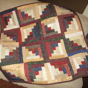May include: A square quilt featuring a log cabin pattern composed of multiple fabric blocks. The design incorporates a variety of colors including deep red, navy blue, forest green, and cream, with intricate swirling patterns stitched throughout.