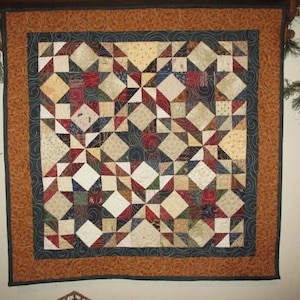 May include: A square wall quilt featuring a patchwork star pattern in shades of cream, navy blue, dark red, and forest green. The quilt is framed by a dark navy blue border with swirling stitching and an outer border of textured rust-orange fabric.