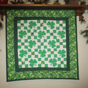 May include: A green and white quilt featuring a shamrock pattern. The quilt has a white background with green shamrocks and green and white checkered squares. The border is green with a floral pattern. The quilt is hanging on a wooden shelf.