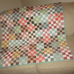 May include: A patchwork quilt with a checkered pattern in various shades of red, pink, brown, green, blue, and white. The quilt is made of squares of different fabrics and is edged with a teal border.