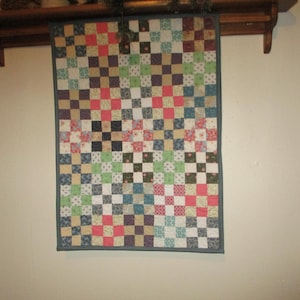May include: A colorful patchwork quilt with a checkered pattern in shades of red, green, blue, and white. The quilt is framed with a dark blue border and is hanging on a wall.