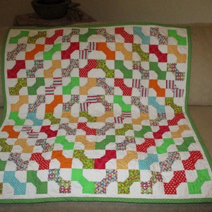 May include: A colorful patchwork quilt with a white background and a repeating pattern of connected shapes in red, green, orange, and blue. The quilt has a green border and is displayed on a beige sofa.