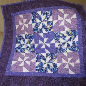 May include: A handmade quilt featuring a pinwheel design in shades of purple, white, and blue. The quilt has a floral pattern in the center and a solid purple border with a swirl pattern. The quilt is approximately 40 inches by 40 inches.