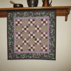 May include: A purple and brown quilted wall hanging with a geometric pattern. The quilt is framed with a dark purple border and a green and purple floral border.