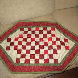 May include: A red and green quilted table runner with a checkered pattern in the center. The runner has a scalloped edge and is made of fabric with a subtle swirl pattern.