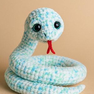 May include: A plush snake toy crafted from soft, variegated yarn in shades of white, light blue, and mint green. The stuffed animal features large, round, dark eyes with teal irises and a bright red, bifurcated felt tongue protruding from its mouth.
