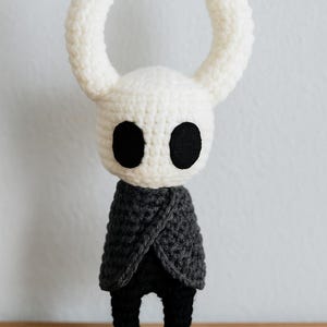 Puede incluir: Mu&ntilde;eca de ganchillo hecha a mano con cabeza blanca, ojos negros grandes y cuernos curvos. La mu&ntilde;eca lleva una capa gris oscuro y pantalones negros. La mu&ntilde;eca se exhibe sobre una superficie de madera.