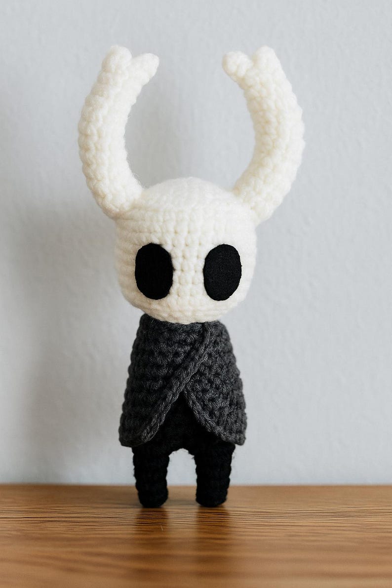 Puede incluir: Mu&ntilde;eca de ganchillo hecha a mano con cabeza blanca, ojos negros grandes y cuernos curvos. La mu&ntilde;eca lleva una capa gris oscuro y pantalones negros. La mu&ntilde;eca se exhibe sobre una superficie de madera.