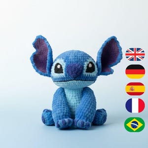 Puede incluir: Una muñeca Stitch azul hecha a mano con ganchillo, con orejas grandes, una nariz azul oscuro y ojos negros. El peluche está sentado sobre una superficie azul claro. Las orejas tienen un interior morado.