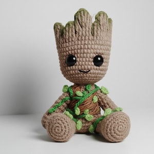 Puede incluir: Muñeco de ganchillo de un personaje con cuerpo marrón, detalles verdes y ojos negros grandes. El muñeco tiene una cara sonriente y está decorado con hojas y enredaderas verdes. El muñeco está sentado.