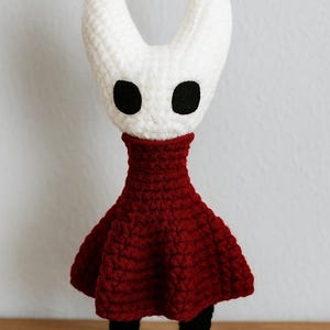 Puede incluir: Mu&ntilde;eca de ganchillo hecha a mano con cabeza blanca, dos cuernos curvos y ojos negros. Tiene un vestido rojo oscuro y piernas negras. La mu&ntilde;eca est&aacute; sobre una superficie de madera.