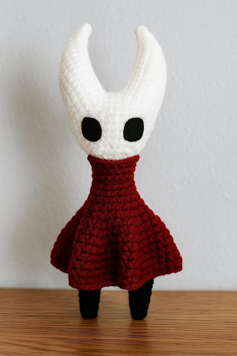 Puede incluir: Mu&ntilde;eca de ganchillo hecha a mano con cabeza blanca, dos cuernos curvos y ojos negros. Tiene un vestido rojo oscuro y piernas negras. La mu&ntilde;eca est&aacute; sobre una superficie de madera.