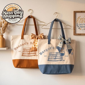 Könnte beinhalten: Zwei Tragetaschen aus Segeltuch hängen an goldenen Kleiderbügeln. Die linke Tasche ist cremefarben mit braunem Boden und zeigt einen Bücherstapel, ein Weinglas und den Text Hannah's Book. Die rechte Tasche ist cremefarben mit blauem Boden und zeigt einen Bücherstapel, ein Weinglas und den Text Margot's Book Club 2026.