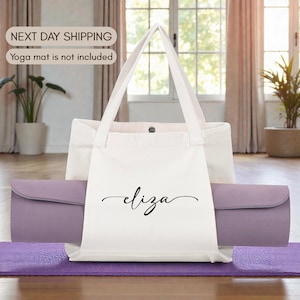 May include: A white canvas tote bag with the name "Eliza" in black script. A rolled-up lavender yoga mat is inside the bag. The bag is on a purple yoga mat. The background includes a window and plants.