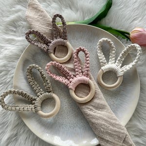 Könnte beinhalten: Vier Makramee-Serviettenringe in Hasenohrenform in Grau-, Rosa- und Weißtönen, jeweils mit einem Holzringsockel. Die Serviettenringe sind auf einem weißen Teller mit einer beigen Serviette angeordnet.
