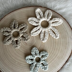Könnte beinhalten: Drei Makramee-Blumendekorationen in Creme-, Salbeigrün- und Beigetönen, angeordnet auf einer Holzscheibe. Die Blumen haben eine kreisförmige Mitte und ein Blütenblattdesign, das eine handwerkliche Ästhetik zeigt. Der Hintergrund ist eine weiche, weiße Oberfläche.