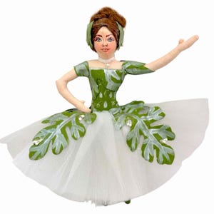 May include: A fabric doll dressed as a ballerina, featuring a green bodice with a white leaf pattern, a white tulle skirt adorned with green leaf appliqués, and brown hair styled in an updo with a green headband and a small white pearl necklace.