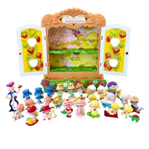 Puede incluir: Un armario de juguete de plástico color canela con dos puertas abatibles que muestran ilustraciones de fresas y el texto The Wonderful World of Strawberry Shortcake, acompañado de una colección de veintidós pequeñas figuras coloridas en varias poses.