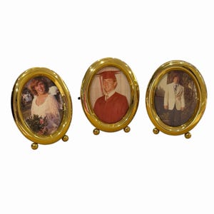 May include: Three oval-shaped gold-toned metal picture frames, each supported by two small spherical feet at the base. The frames contain vintage photographs: one showing a person in a white dress, one a graduate in a cap and gown, and one in a suit.