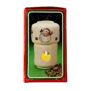 May include: A white ceramic oil warmer featuring a Santa Claus illustration with holly accents. The cylindrical base has a circular cutout revealing a lit tea light candle inside. The top bowl is designed to hold oil or wax for fragrance diffusion.