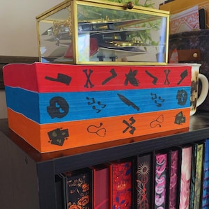 Op de afbeelding: Een handgeschilderde opbergdoos met drie niveaus en rode, blauwe en oranje secties. Elke sectie bevat zwarte illustraties van schedels, messen en andere symbolen. Bovenop staat een goudkleurige glazen doos en daaronder een boekenplank.