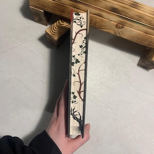 May include: A decorative book with a spine featuring a botanical design of vines and leaves in green and brown. The book's edge is red, and a black hand graphic is visible. The book is held in a person's hand.