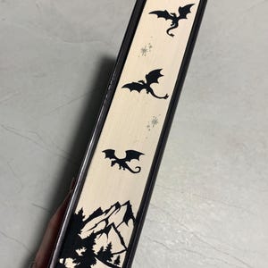 Puede incluir: Un libro negro y crema con temática de fantasía. El lomo presenta tres siluetas de dragones negros y una escena de montaña y bosque. El libro tiene un borde negro.