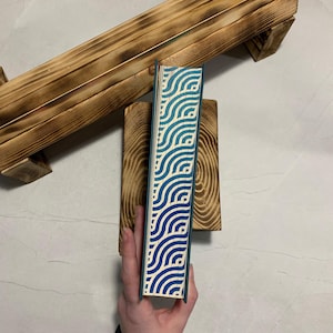 Puede incluir: Una bandeja de baño de madera con acabado quemado, un pequeño bloque de madera y un libro con un patrón de olas azul y blanco. El libro está en manos de una persona.