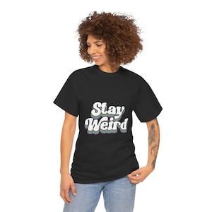 Puede incluir: Camiseta negra con el texto "Stay Weird" en una fuente retro. El texto es blanco con contornos verdes, morados y azules. La camiseta la lleva una persona con pelo castaño rizado y vaqueros azules.