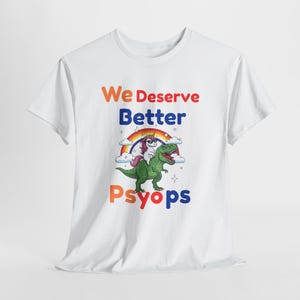 May include: White t-shirt with the text "We Deserve Better Psyops" in orange, blue, and red. A cartoon illustration features a unicorn riding a green dinosaur under a rainbow.
