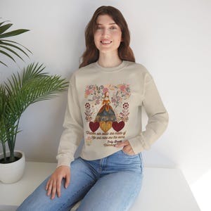 May include: Cream-coloured sweatshirt with a decorative illustration of a woman in a dress, floral elements, and hearts. The text "Whatever our souls are made of, His and mine are the same" is printed below the illustration. The sweatshirt is worn by a person.