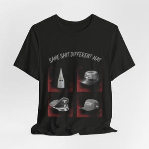 以下が含まれることがあります： グレーのテキスト「SAME SHIT DIFFERENT HAT」がプリントされた黒の半袖Tシャツ。テキストの下には、赤くかすれた背景の4つの正方形パネルがあり、それぞれ白いフード、グレーのミリタリーキャップ、ダークな制帽、グレーの野球帽が描かれています。