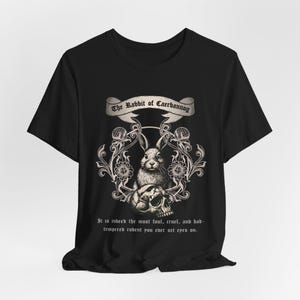 Op de afbeelding: Zwart T-shirt met een gedetailleerde illustratie van een konijn, schedel en bloemelementen. De tekst "The Rabbit of Caerbannog" staat boven het ontwerp, met extra tekst eronder. Een uniek grafisch T-shirt.