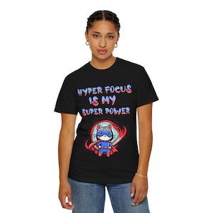 Op de afbeelding: Zwart T-shirt met de tekst "HYPER FOCUS IS MY SUPER POWER" in blauw en rood. Een cartoonkat in een superheldenpak staat onder de tekst. De kat draagt een masker en een cape.