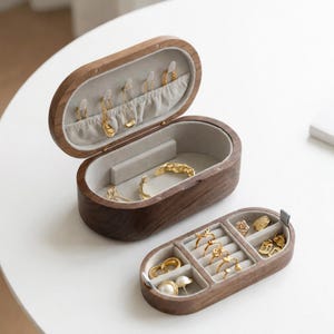 Könnte beinhalten: Zwei ovale Schmuckkästchen aus Holz. Die größere Box ist geöffnet und zeigt einen grauen Innenraum mit Haken für Halsketten. Die kleinere Box ist geöffnet und zeigt Ringe und Ohrringe. Goldschmuck ist sichtbar.