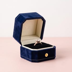 May include: A navy blue velvet ring box with an octagonal shape and white stitching. The box is open, revealing a cream-colored interior lining and a black velvet insert holding a rose gold ring with a clear, faceted, round-cut center stone.