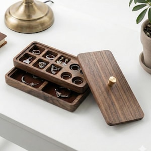 May include: A wooden jewelry organizer with multiple compartments, holding rings, earrings, and bracelets. The organizer is open, revealing the jewelry inside. The lid is off to the side, showing a gold-colored knob. The organizer is on a white surface.