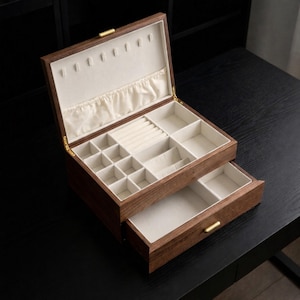 Puede incluir: Caja de joyería de madera marrón oscuro con tapa con bisagras y cajón extraíble. El interior claro tiene varios compartimentos para guardar joyas. La ferretería es de color dorado.