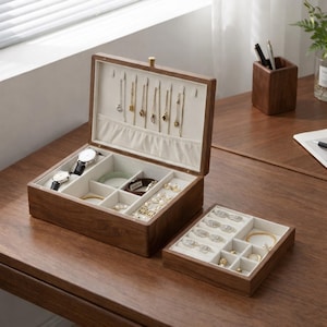 Könnte beinhalten: Eine Schmuckschatulle aus dunklem Holz mit offenem Deckel, die Fächer für Uhren, Armbänder und Halsketten zeigt. Die Box hat eine hellfarbene Innenausstattung. Daneben befindet sich ein kleineres, passendes Tablett, ebenfalls mit Schmuck gefüllt.