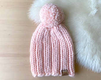 May include: A pink knitted beanie with a fluffy pom pom on top. The beanie has a ribbed texture and a small brown leather tag with the word "Cozy" on it.
