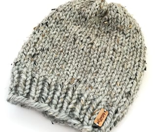 May include: A gray knit beanie with a ribbed design. The hat is made of thick yarn with flecks of black, brown, and white. A small wooden tag with the text "Canopy Knits Co." is attached to the brim.